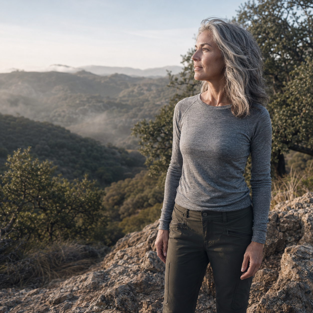 Woman in her early 50s with silvering shoulder-length hair, wearing a heather-gray long-sleeve and olive technical pants, standing at a rocky Topanga Canyon overlook in soft morning light, looking thoughtfully across the chaparral hillside, representing the mental clarity and healthy brain aging support associated with Cerebrolysin at Evolve Anti-Aging & Wellness in Encino, CA