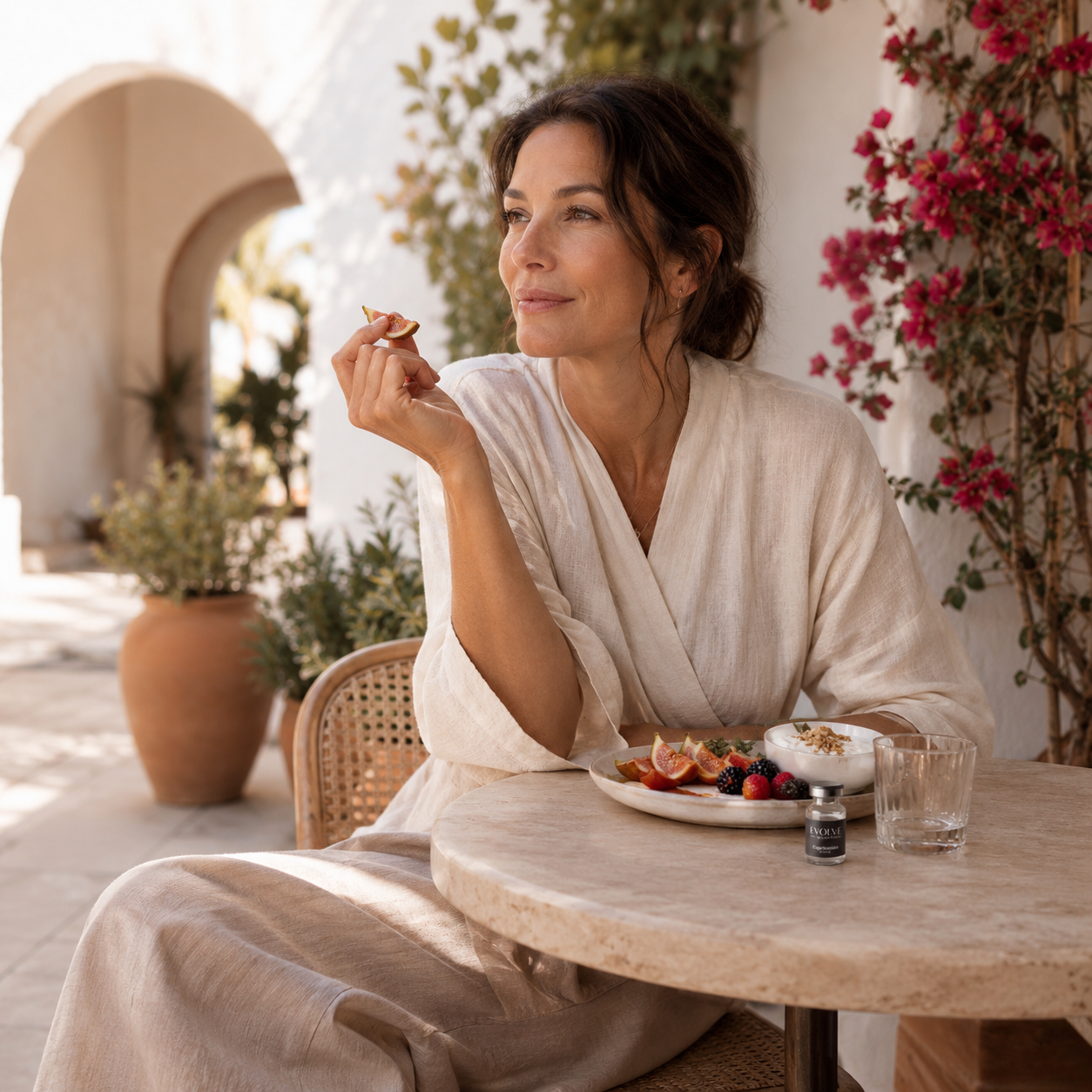 Woman in her mid-40s wearing a cream linen wrap top, seated at a travertine bistro table in a sunlit Mediterranean garden courtyard with bougainvillea and terracotta planters, enjoying a small Mediterranean breakfast of fresh figs, berries, and yogurt, EVOLVE Cagrilintide peptide vial discreetly placed on the table, representing the mindful wellness and healthy body composition support associated with Cagrilintide at Evolve Anti-Aging & Wellness in Encino, CA
